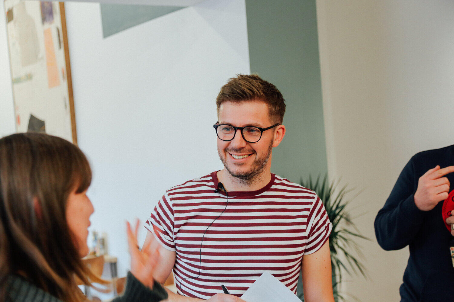 Content designer, Iain Broome from Very Meta Iain Broome in a striped t-shirt, smiling as he talks to a member of the audience at Sheffield Content Club. There is a white wall behind him with the leaves of some lovely indoor plants in view too.